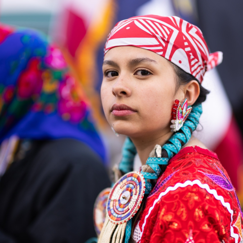 Young woman in traditional attire and beaded jewelry, looking at the camera outdoors.