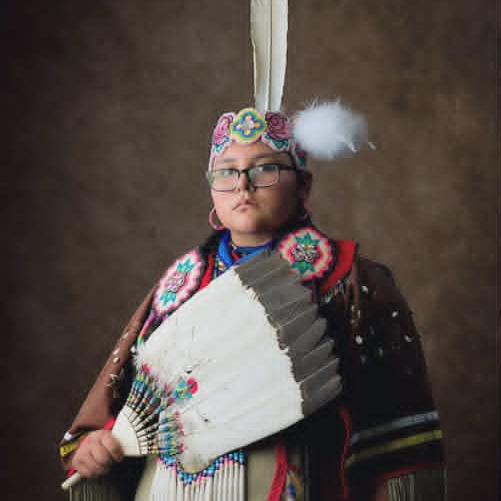 Person in traditional Native regalia holding a feather fan, standing against a brown backdrop.