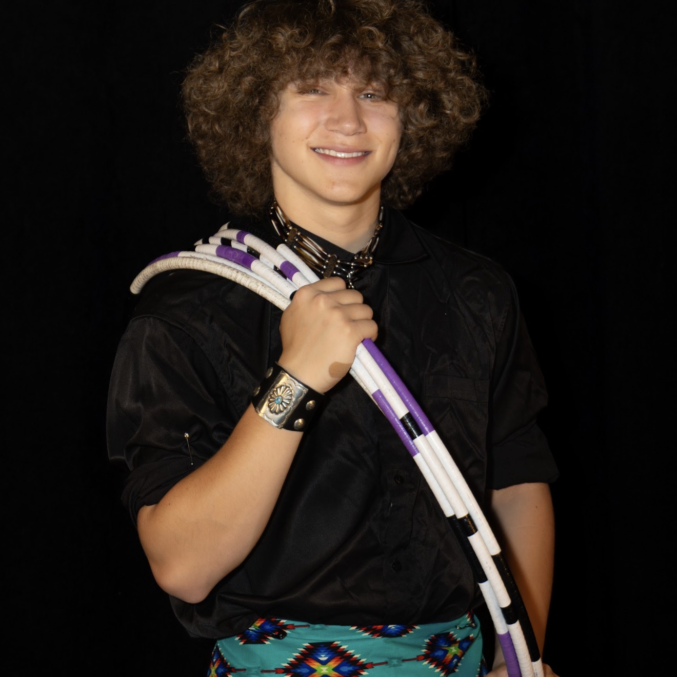 Smiling person in beadwork skirt holding hoops, wearing a black shirt and beaded necklace.
